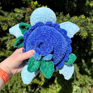May include: A handmade plush flower in shades of blue and green. The central bloom is a deep blue, surrounded by lighter blue petals and green leaves. The plush toy is soft and textured, with a round shape.