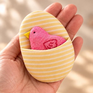 May include: A small, oval-shaped fabric pouch featuring a yellow and white horizontal striped pattern. Tucked inside the pouch is a bright pink plush bird ornament with a small yellow beak and a stitched wing detail on its side.