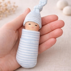 May include: A handmade baby doll with a wooden head and painted facial features. The doll is wrapped in blue and white striped fabric and wears a matching cap. The doll is held in a hand.