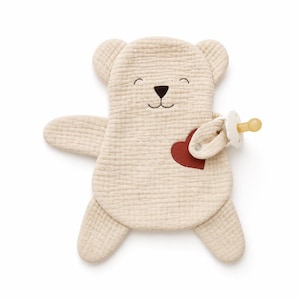 Puede incluir: Un peluche de oso de muselina beige con tela texturizada, cara bordada en marrón oscuro y un parche de corazón rojo oscuro en el pecho. Un chupete blanco está sujeto al brazo del oso con una pequeña correa de tela y un broche de metal plateado.