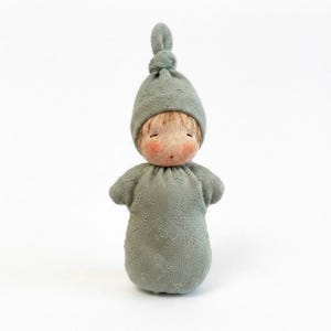 Puede incluir: Una pequeña muñeca con cara redonda, mejillas sonrosadas y cabello castaño claro, que lleva un gorro de punto verde salvia con un nudo superior y un arrullo de punto verde salvia a juego con un sutil patrón floral bordado en tono.