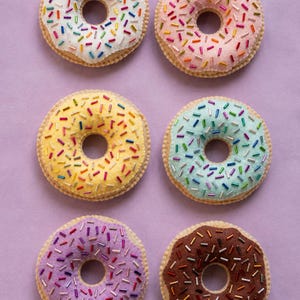 Puede incluir: Seis juguetes de fieltro con forma de dónut dispuestos en dos columnas. Cada dónut presenta un glaseado de distinto color—blanco, rosa, amarillo, azul claro, morado y marrón—decorado con pequeñas virutas cilíndricas de colores variados.