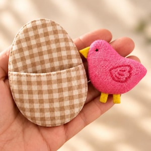Op de afbeelding: Een eivormig stoffen zakje met een beige en wit vichy-ruitpatroon, gecombineerd met een kleine, felroze knuffelvogel met een gele snavel, gele pootjes, één zwart oogje en een geborduurd vleugeldetail aan de zijkant.