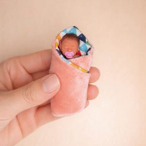 May include: A miniature baby doll wrapped in a pink blanket. The baby has brown skin, closed eyes, and a pink pacifier. The blanket has a colorful patterned lining with blue, white, and yellow accents.