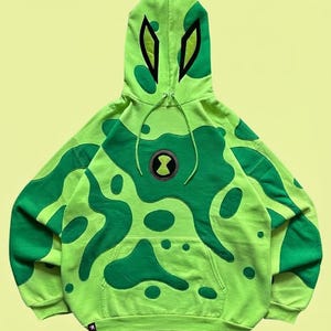 May include: A lime green hoodie featuring dark green organic-shaped patches throughout the body and sleeves. The hood displays two black-outlined eye shapes, and the chest features a circular black and green emblem with a central hourglass symbol.