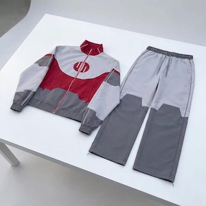 Könnte beinhalten: Ein zweiteiliger Trainingsanzug in Grau, Rot und Weiß. Die Jacke hat einen hohen Kragen, eine rote Brustpartie und einen Reißverschluss. Die Hose ist weit geschnitten mit einem Kordelzug in der Taille und grauen Einsätzen am unteren Saum.