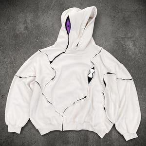 Può includere: Felpa con cappuccio bianca con un design unico caratterizzato da linee nere e un dettaglio occhio viola sul cappuccio. Un emblema in bianco e nero è sulla parte anteriore. La felpa ha una vestibilità ampia e maniche lunghe.