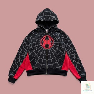 May include: A black zip-up hoodie featuring a grey spiderweb pattern across the front, back, and sleeves. A large red spider emblem is centered on the chest. The garment includes red side panels, a hood, and a full-length silver metal zipper.