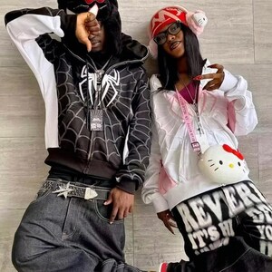 May include: A black zip-up hoodie with a white spiderweb graphic and a white spider logo, paired with dark gray denim jeans and a star-buckle belt. Beside it, a white zip-up hoodie with pink accents, black graphic pants, and a round Hello Kitty bag.