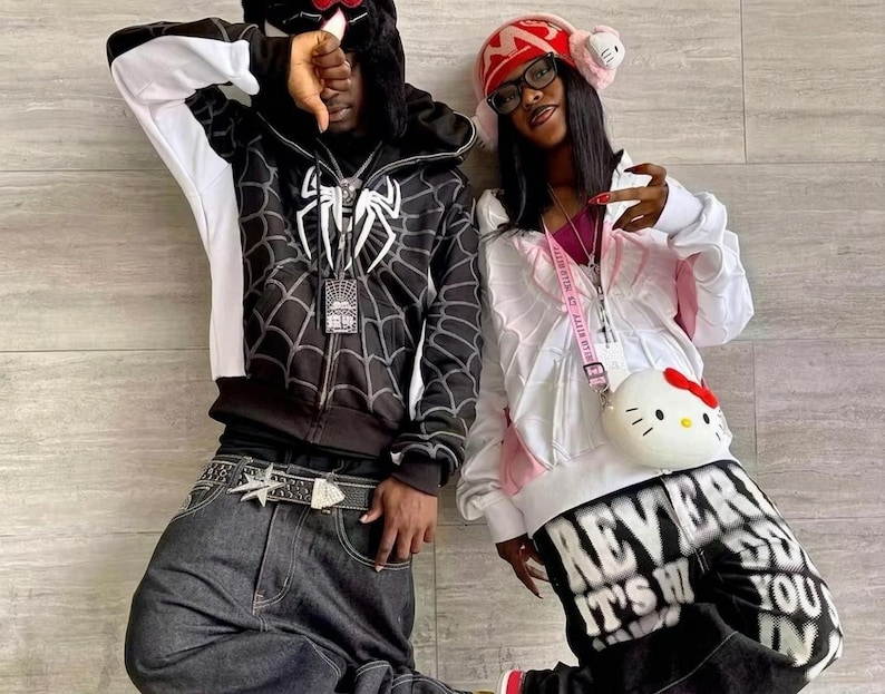 May include: A black zip-up hoodie with a white spiderweb graphic and a white spider logo, paired with dark gray denim jeans and a star-buckle belt. Beside it, a white zip-up hoodie with pink accents, black graphic pants, and a round Hello Kitty bag.