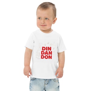 May include: A white short-sleeved t-shirt featuring a red graphic print of the text DIN DAN DON stacked vertically. To the left of the text is a thin-lined illustration of a vintage gramophone with musical notes floating above it.