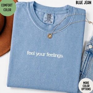 May include: A blue jean-colored t-shirt with the phrase "feel your feelings" in white script. A gold-colored necklace with a pendant is draped across the shirt. The shirt is folded, and the words "Comfort Color" are visible.