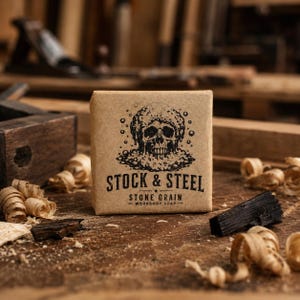 May include: A square bar of soap wrapped in brown paper with a black ink illustration of a skull surrounded by bubbles. The text reads STOCK & STEEL, STONE GRAIN, and WORKSHOP SOAP. The design is centered on the front of the paper packaging.