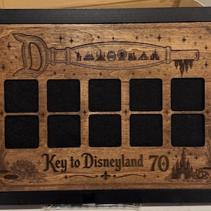 Könnte beinhalten: Holz-Schattenbox mit schwarzem Rahmen. Die Box zeigt ein Schlüsseldesign mit Disneyland-Symbolen und dem Text "Key to Disneyland 70". Sie hat zehn quadratische Öffnungen mit schwarzem Stoffrücken.