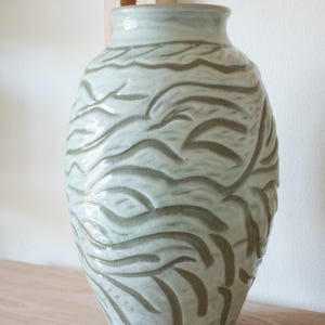 Könnte beinhalten: Eine hellgrüne Keramikvase mit glänzender Oberfläche und strukturierter Textur, die mit geschnitzten, wellenförmigen, horizontalen Linien verziert ist. Die Vase hat eine bauchige Form mit einem schmalen Hals und einem glatten Rand.