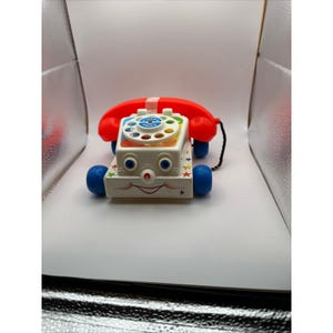 Può includere: Un telefono giocattolo vintage Fisher-Price con una cornetta rossa e una base bianca. La base ha ruote blu, una faccia sorridente e decorazioni a stella. Il quadrante è bianco con dettagli blu e verdi.
