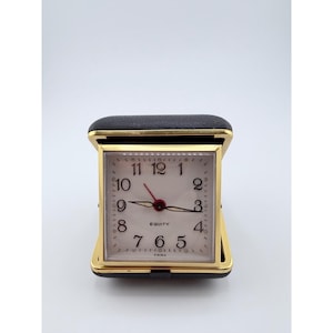 Puede incluir: Un reloj despertador de viaje cuadrado con marco de metal dorado y estuche plegable de cuero sintético negro. La esfera blanca presenta números arábigos negros, manecillas de hora y minutos negras, un segundero rojo y el texto Equity y China.
