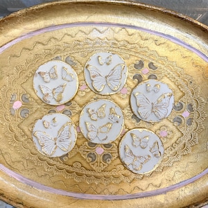 May include: Six circular white clay discs featuring raised butterfly motifs with gold leaf accents, arranged on an oval gold-toned decorative tray with intricate embossed patterns and small pink painted details.