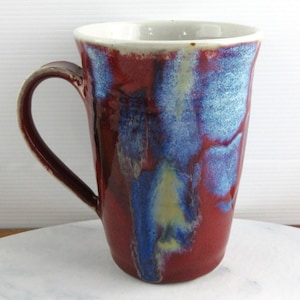 May include: A handcrafted ceramic mug with a distinctive glaze. The mug has a deep red base with blue and yellow streaks, and a white interior. It features a curved handle and is suitable for hot or cold drinks.