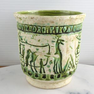May include: A cylindrical ceramic planter with a cream-coloured base and a textured green glaze. The exterior features embossed relief carvings of stylised horses and geometric patterns encircling the rim and base, with a smooth green interior.