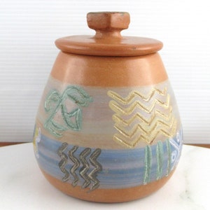 May include: A terracotta ceramic jar with a lid, decorated with a geometric pattern in shades of blue, green, and gold. The jar has a rounded shape and a slightly flared opening.