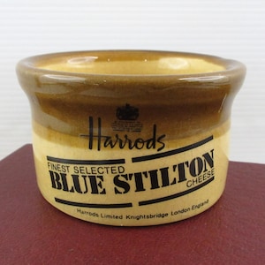 May include: A small, round ceramic container with a brown and tan glaze. The container features the Harrods logo and the text "FINEST SELECTED BLUE STILTON CHEESE". The text is in a bold, black font.