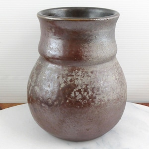 May include: A handmade, brown ceramic vase with a textured finish. The vase features a rounded body and a gently flared rim. The interior is a darker brown. The vase is presented on a white marble surface.