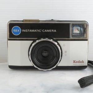 以下が含まれることがあります： ヴィンテージのKodak Instamatic 155Xカメラ。カメラは黒い上部と大きなレンズを備えた白いボディをしています。「155X Instamatic Camera」の文字が前面に印刷されています。黒いリストストラップが付いています。イギリス製。