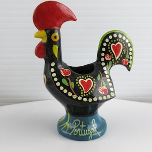 May include: A black ceramic rooster figurine with red, yellow, and green details. The base is blue and inscribed with "Portugal". The rooster features a red comb and wattle, with heart and floral patterns. The figurine is a decorative item.