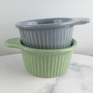 May include: Two small, stacked ceramic ramekins. The bottom ramekin is light green, and the top one is grey. Both have a ribbed design and a single handle. They are sitting on a white marble surface.