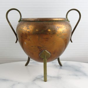 Puede incluir: Un recipiente redondo de cobre con dos asas curvas en los lados opuestos y tres patas de latón con fijaciones en forma de hoja en la base. La superficie de cobre muestra una pátina desgastada con variaciones visibles de color y textura.