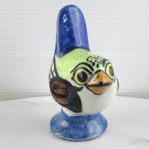 Puede incluir: Una pequeña figurita de pájaro de cerámica con un diseño pintado en azul, verde y blanco. El pájaro tiene una cabeza grande y redonda con un pico amarillo y ojos negros. El pájaro está de pie sobre una base azul.