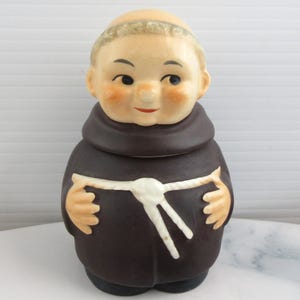 May include: A ceramic figurine of a monk, featuring a brown robe and a white rope belt. The monk has a bald head, rosy cheeks, and a smiling expression. The figurine is a decorative item.