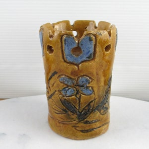 May include: A small, brown ceramic candle holder with blue floral designs. The holder has a decorative, openwork design around the top.