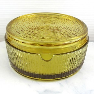 May include: A vintage, yellow-green glass ashtray with a lid. The lid has a textured, concentric circle design. The ashtray's base has a vertical ribbed pattern. The ashtray is sitting on a white surface.