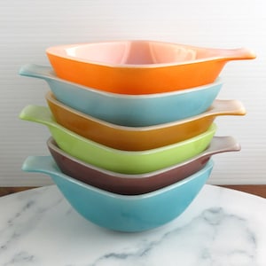 Puede incluir: Una pila de seis cuencos de vidrio vintage coloridos. Los cuencos son en tonos naranja, azul, verde, marrón y turquesa. Cada cuenco tiene un asa. Los cuencos están apilados sobre una superficie de mármol blanco.