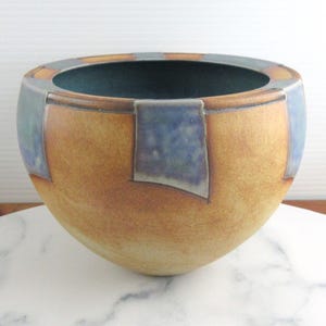 May include: A handcrafted ceramic bowl with a rounded shape. The bowl features a warm, earthy colour palette with a tan base and blue and orange accents. The interior is a deep teal. The bowl is displayed on a white marble surface.