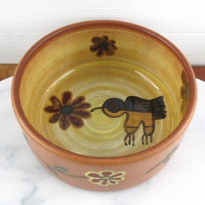 May include: A round ceramic bowl with a brown rim and a yellow interior. The bowl features a hand-painted design of a bird and flowers in brown and black. The bowl is sitting on a white marble surface.