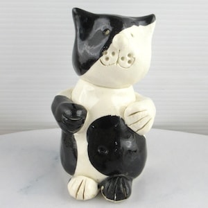 May include: A black and white ceramic cat figurine with a whimsical expression. The cat has a black face and a white body with black spots.