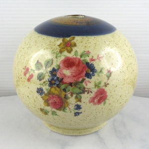 May include: A spherical, cream-coloured ceramic vase with a floral design. The vase has a dark blue band at the top and is decorated with pink roses, blue and yellow flowers, and green leaves. The vase has a speckled texture.