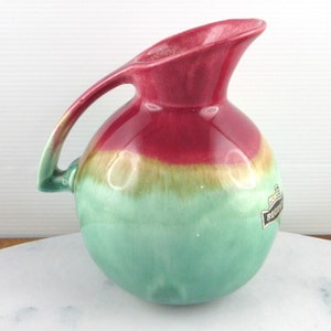 Puede incluir: Una pequeña jarra de cerámica redonda de color verde y rosa con asa. La jarra tiene un esmalte que crea un efecto degradado de rosa a verde. La jarra tiene una etiqueta en el lateral que dice "Regina."