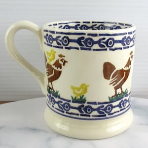 Könnte beinhalten: Cremefarbene Keramiktasse mit Henkel, verziert mit braunen und gelben Hahn- und Kükenillustrationen. Die Tasse hat einen blauen Zierrand und ist für Heißgetränke geeignet. Eine charmante Ergänzung für jede Küche.