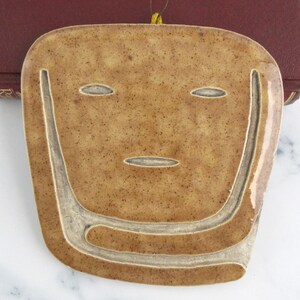 May include: A tan, speckled ceramic wall plaque shaped like a stylized face with incised lines forming the eyes, mouth, and a U-shaped jawline. The piece features a glossy finish and includes a thin, dark string loop at the top for hanging.