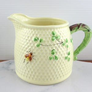 May include: A pale yellow ceramic jug with a textured surface and a green bamboo-style handle. The jug is decorated with green shamrocks and a small insect illustration. The jug is likely for serving beverages.
