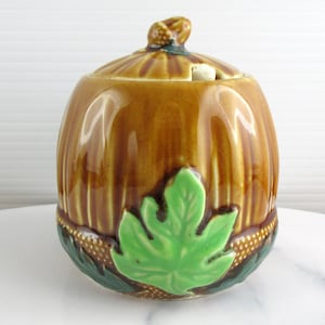 May include: A brown ceramic pumpkin-shaped jar with a lid and a green leaf design. The jar has a textured surface and a small knob on top. The base of the jar has a decorative border.