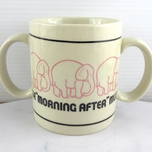 May include: Cream ceramic mug with two handles, decorated with pink elephant outlines and the black text "THE MORNING AFTER". The mug has black bands near the top and bottom.