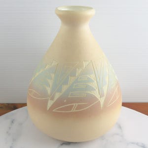 May include: A beige ceramic vase with a rounded body and a narrow neck. The vase features etched geometric designs in shades of blue and white, with a band of pink near the base. The vase is displayed on a marble surface.