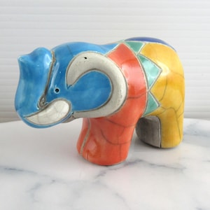 Könnte beinhalten: Eine farbenfrohe Elefantenfigur aus Keramik. Der Elefant ist mit blauen, orangefarbenen und gelben Abschnitten bemalt, mit weißen Akzenten an Rüssel und Stoßzähnen. Die Figur hat ein geometrisches Design und ist ein dekorativer Gegenstand.