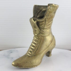 May include: A vintage brass boot figurine. The boot has a pointed toe, a lace-up front, and a small heel. The boot is a golden color and appears to be made of metal. The boot is a decorative item.