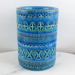 May include: A cylindrical ceramic vase featuring a vibrant blue glaze with horizontal bands of embossed geometric patterns, including rows of small circles, vertical lines, and diamond shapes highlighted with subtle green and teal undertones.